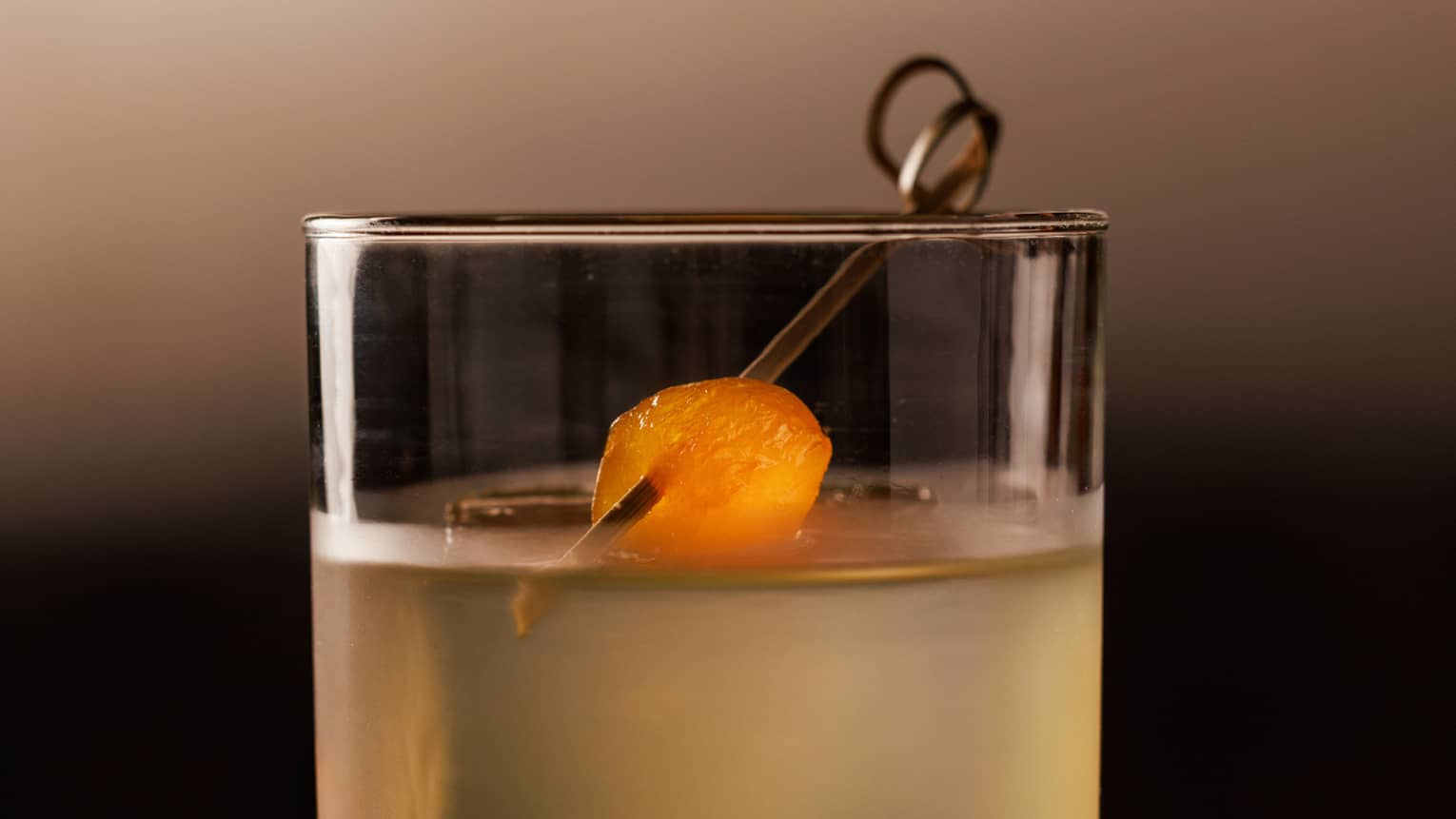 Yellow-coloured cocktail garnished with a piece of papaya on a toothpick, served in a rocks glass