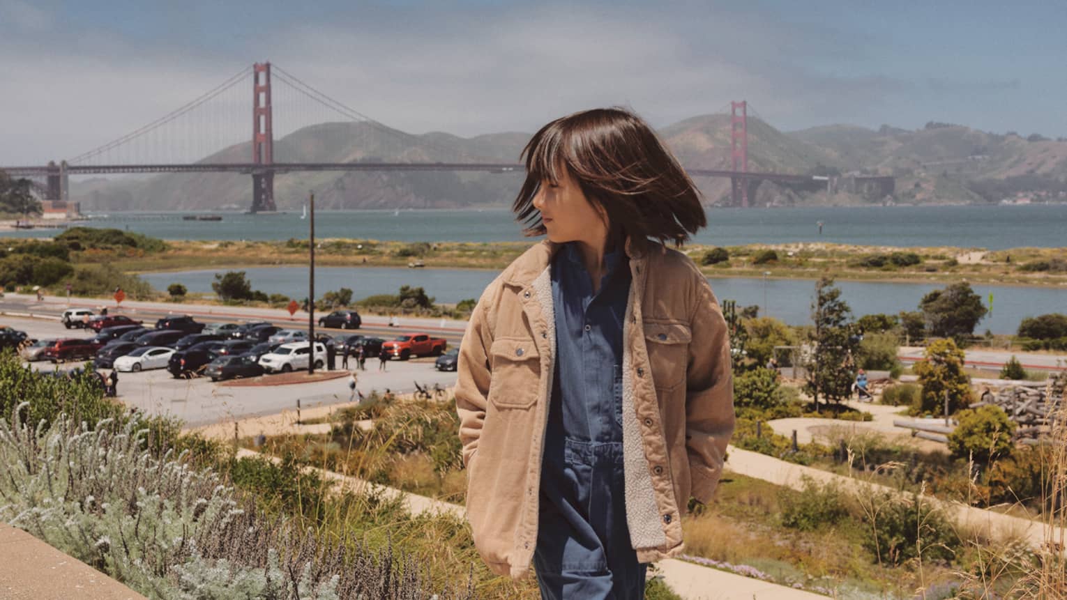 A person with short brown hair wearing a denim jumpsuit and a tan coat walks toward the camera with the Golden Gate Bridge in the background.