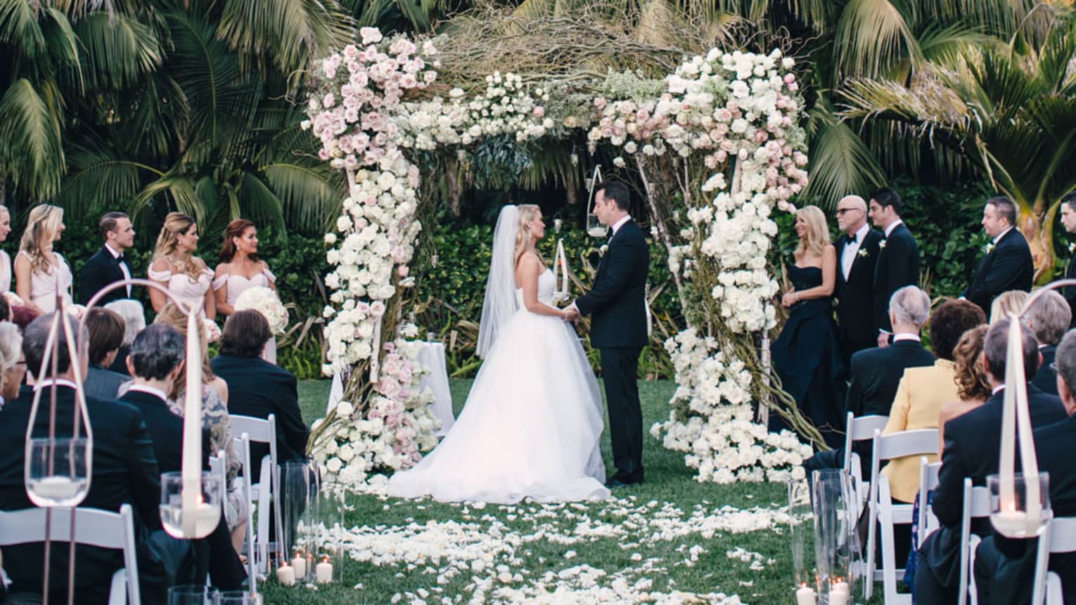 Garden wedding ceremony, bride and groom under rose-covered altar and palm trees