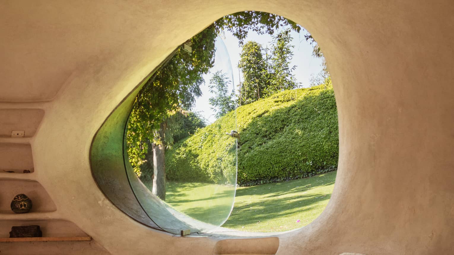 Smooth, adobe wall frames a curved, asymmetrical window, glass pivoted out, a verdant yard with large pruned hedge in view.