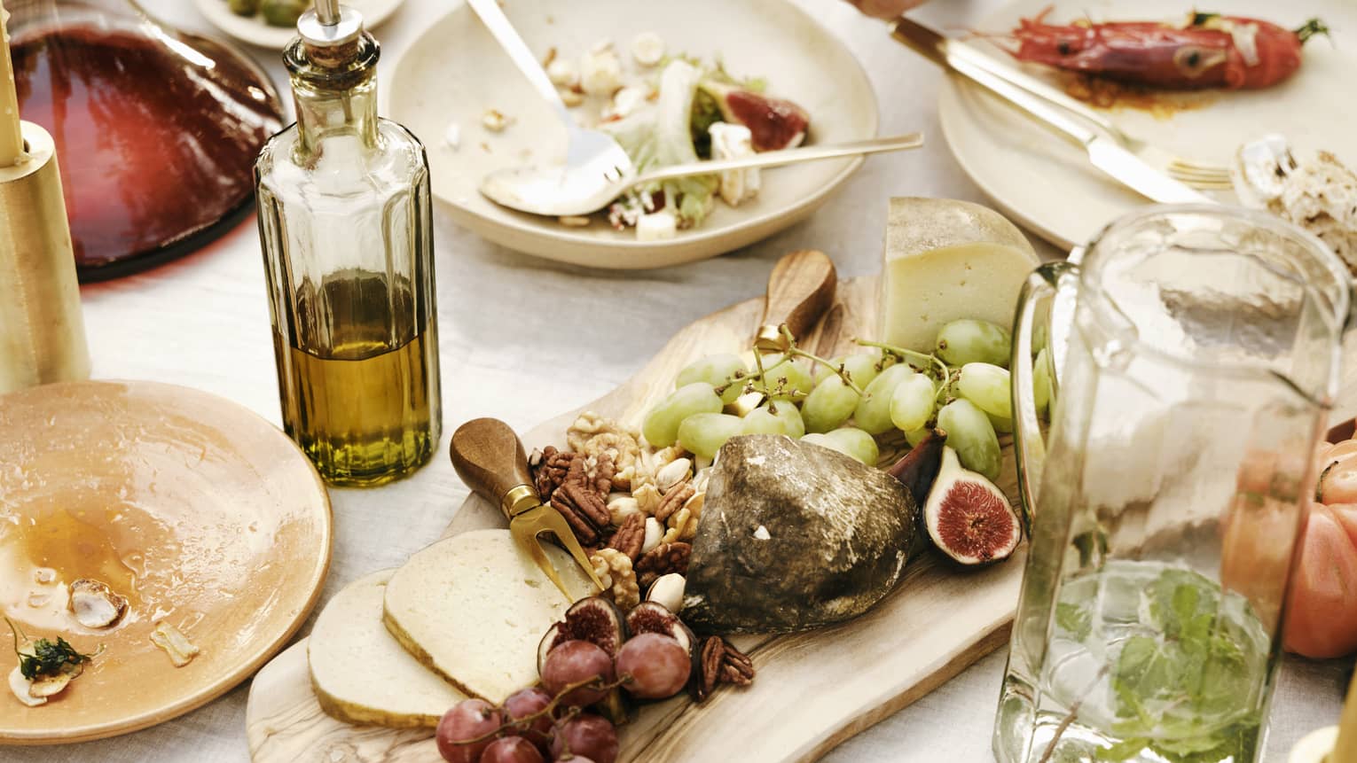 Close-up of a table laden with dishes of fresh figs, grapes, pecans, cheese, seafood, olive oil and a carafe of mint water.