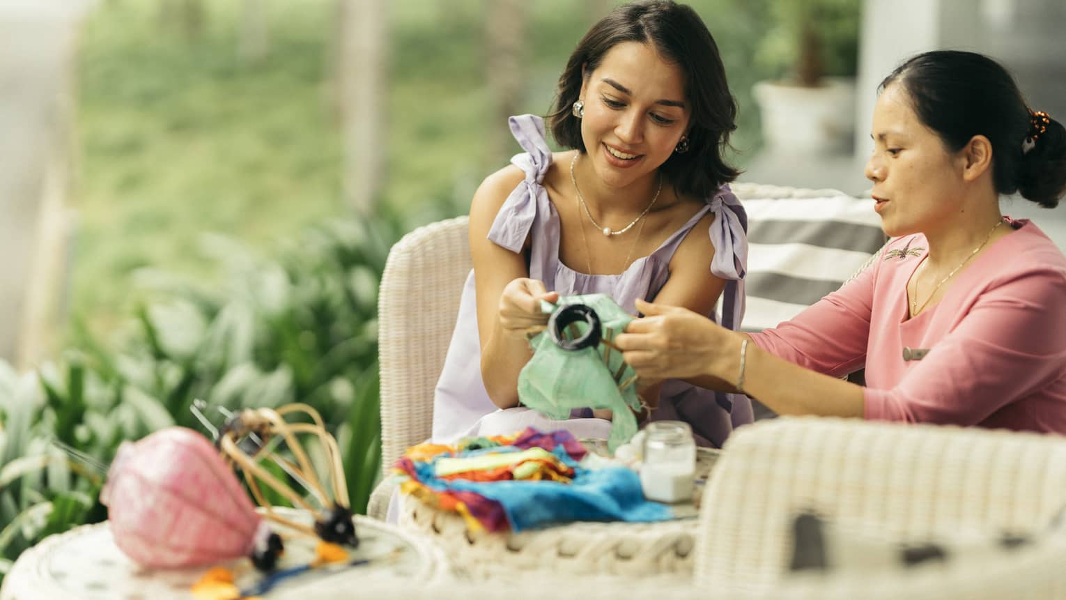 Two people engaged in a traditional craft activity, working together on fabric art while seated on a wicker patio set in a lush, green outdoor setting.