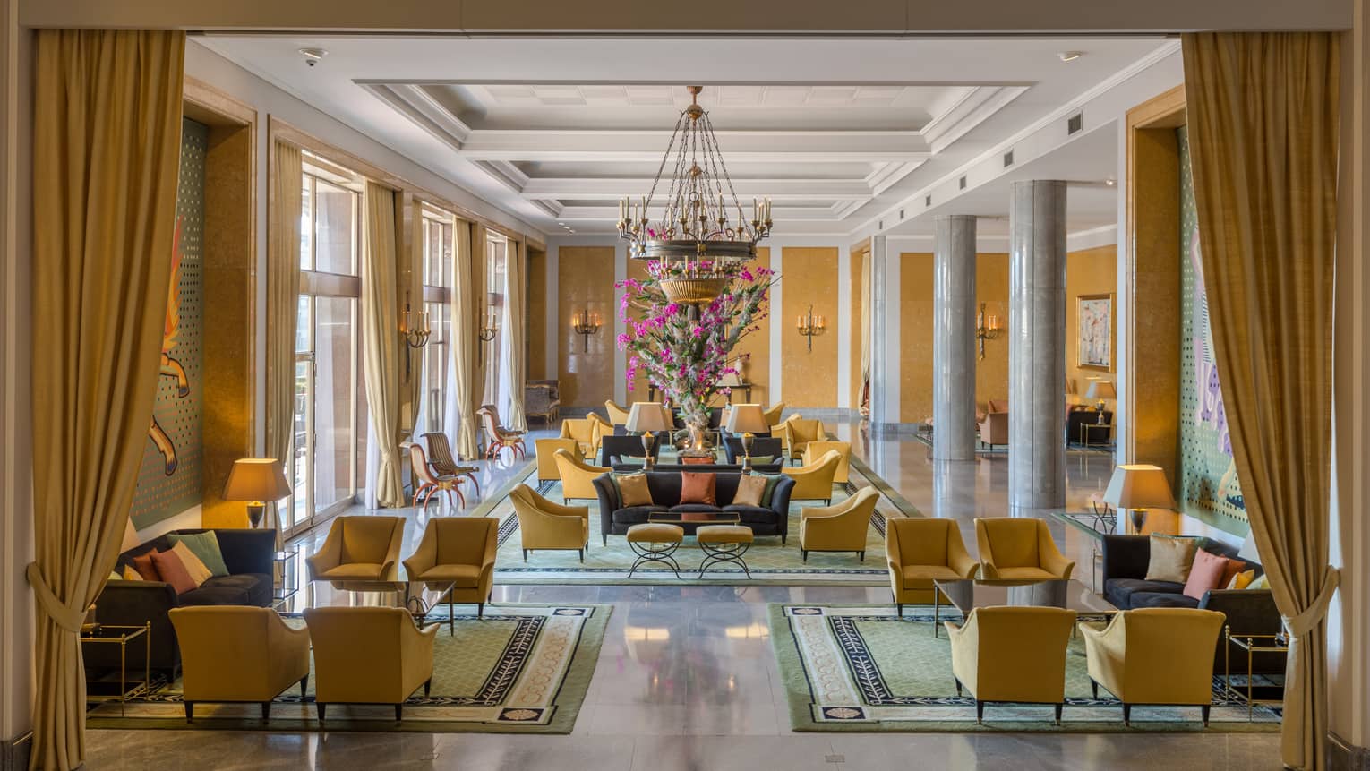 Bright hotel lobby with long gold curtains and armchairs, seating area under chandelier