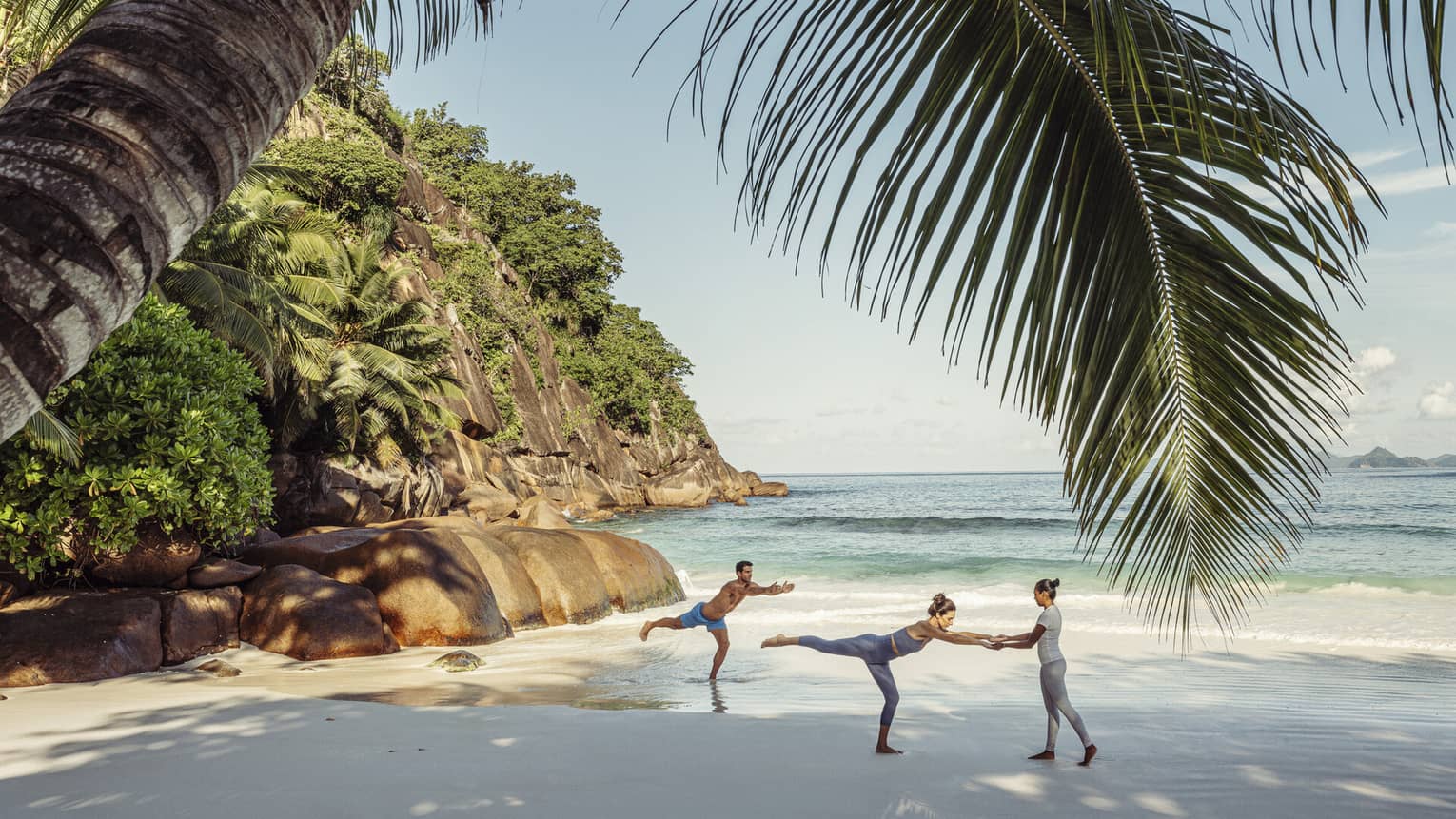 Two guests and an instructor doing yoga on a beach shaded by a large palm tree