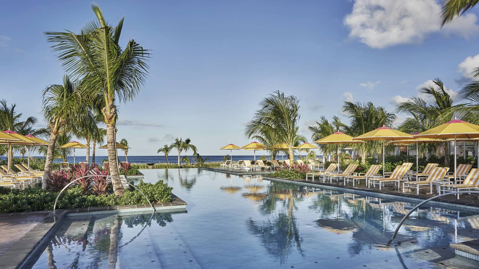 A calm pool reflecting the blue sky and palm trees, lined with lounge chairs and yellow umbrellas