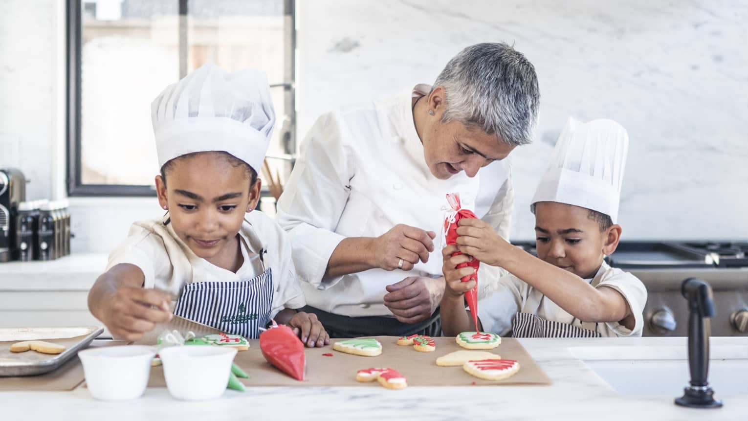 Two children in chef’s hats and aprons in a stark, white kitchen; a chef watches closely as one pipes icing onto a cookie.