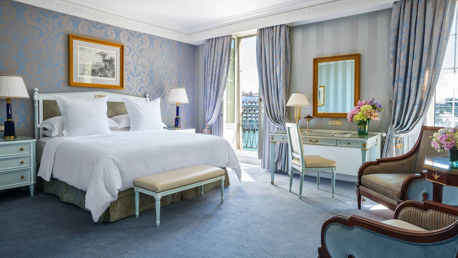 Luxury hotel room with a king bed, nightstands with lamps, seating area, desk and a balcony with light blue and gold decor
