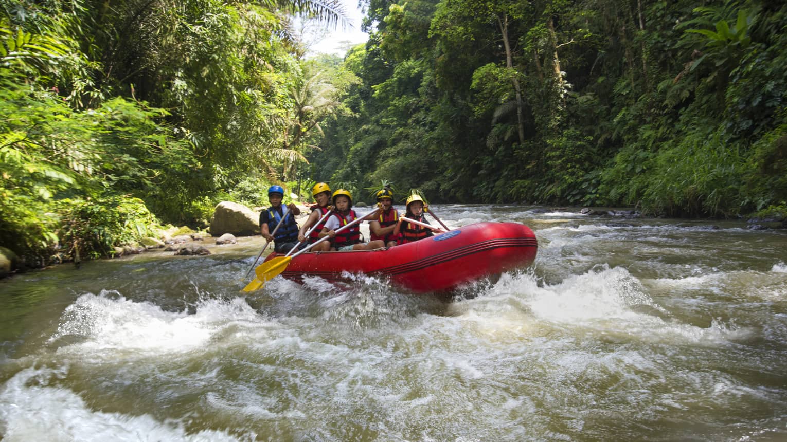 Family of four and guide hold paddles, steer red inflatable raft on rapids in river