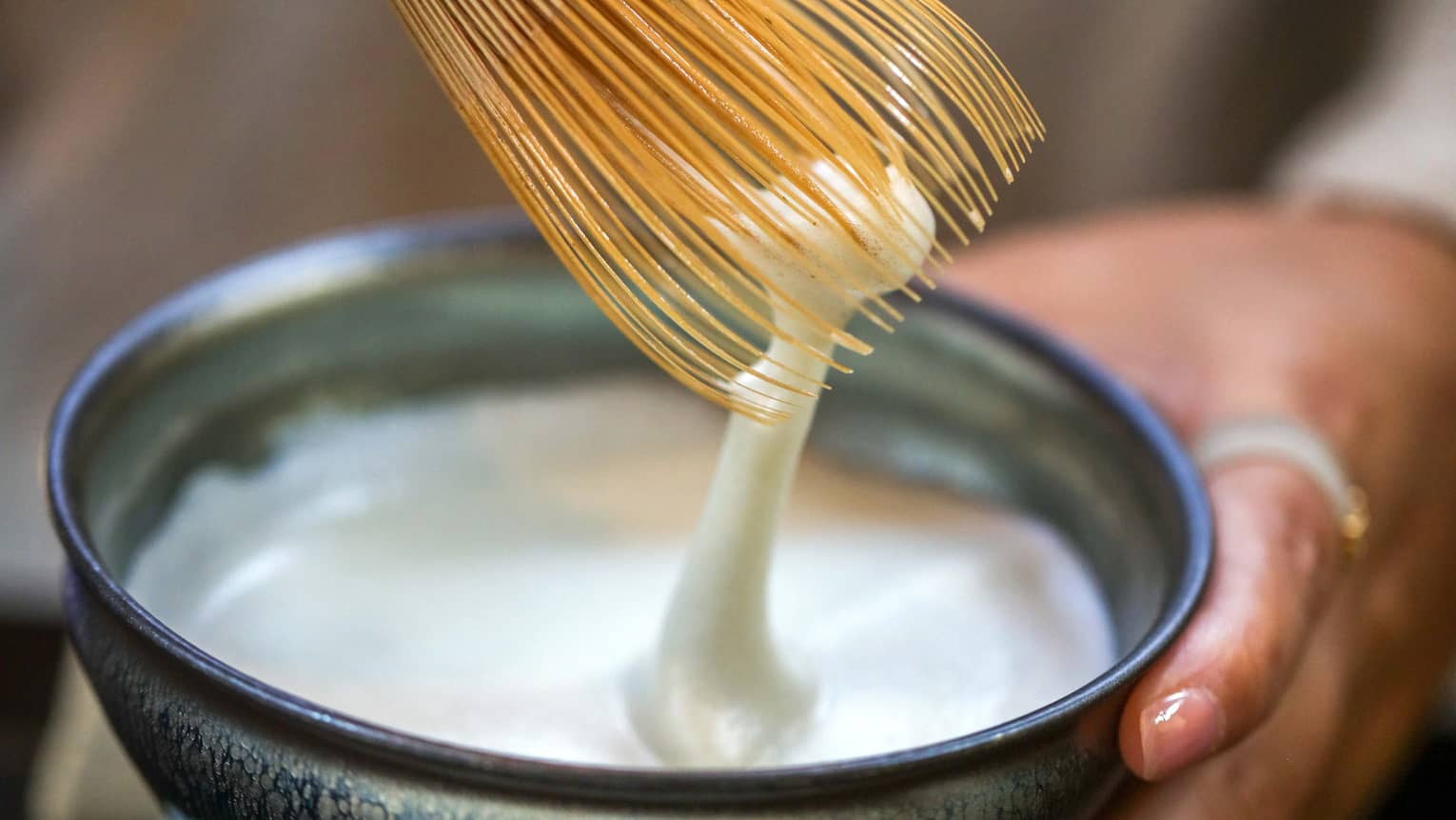 A white foamy liquid being stirred with a straw whisk in a small blue stoneware bowl on a petal-shaped base.