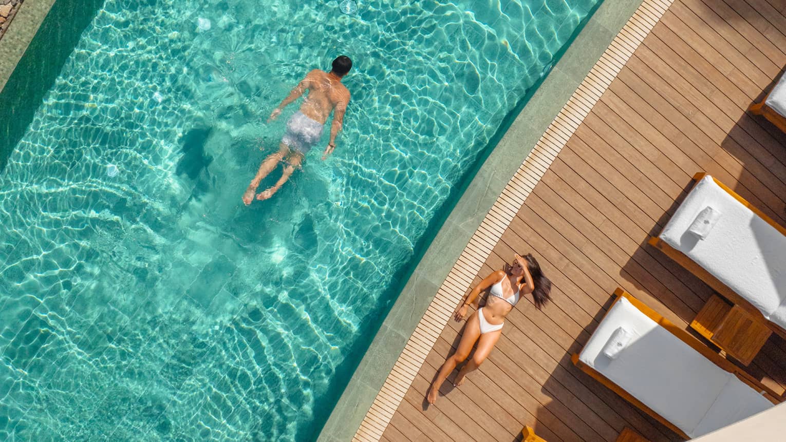 Aerial view of man swimming in a narrow pool with a woman in a white bikini laying next to the pool on a wooden pool deck