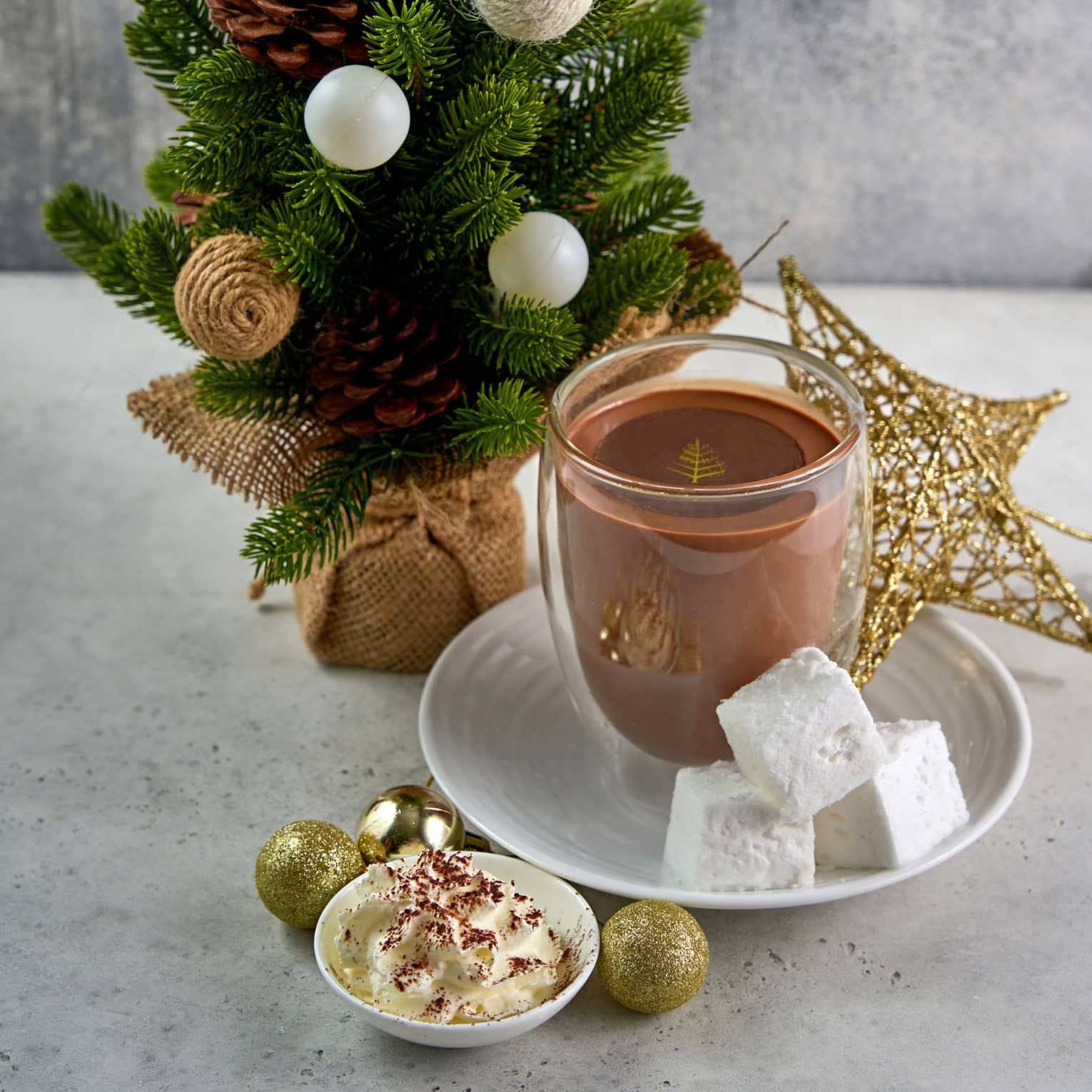 Hot chocolate served in a clear mug with three cubed marshmallows next to it on a plate, set next to a small decorative Christmas tree