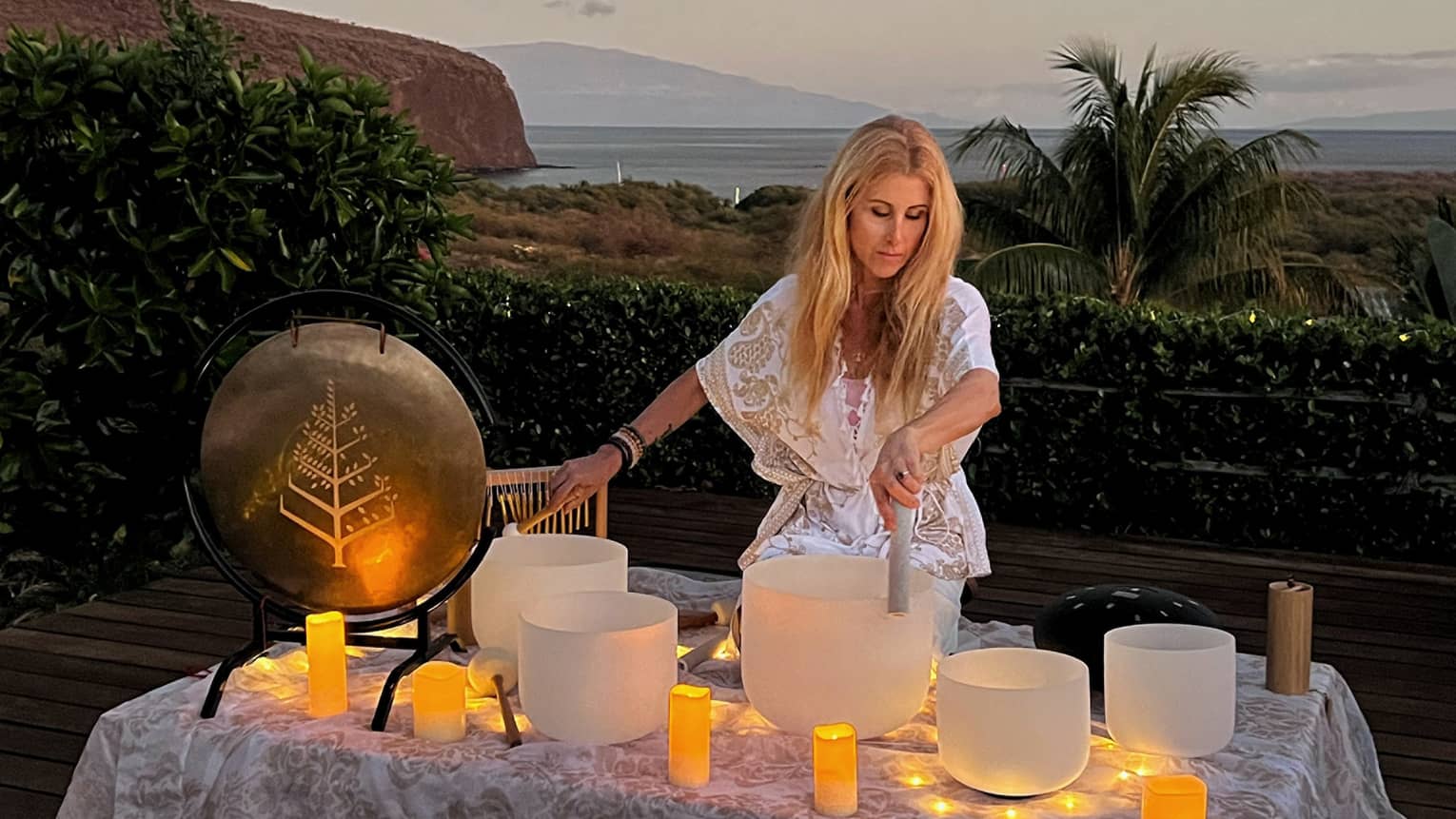 A woman performs crystal singing bowl ritual outside in beach setting at dusk