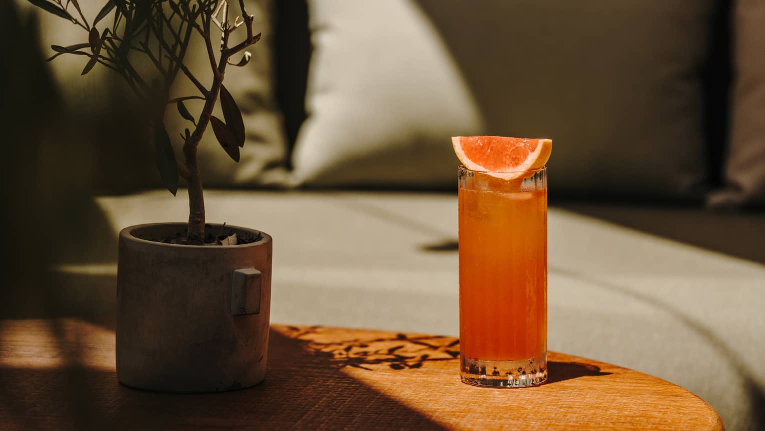 A small round table holds a tall glass of a pinkish-orange drink garnished with citrus.