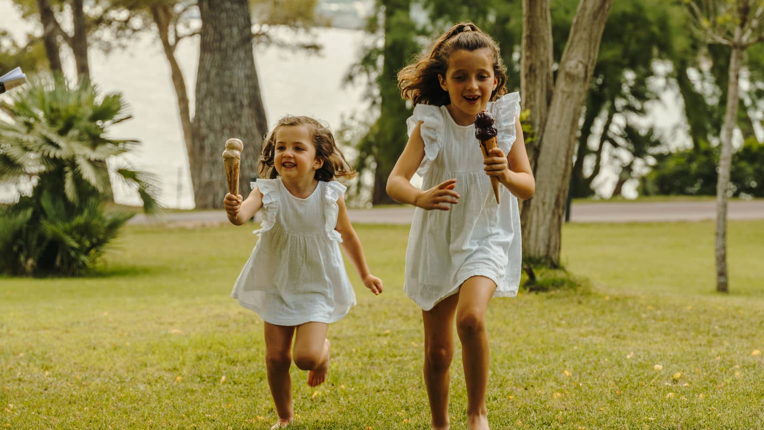 Two children holding double-scoop ice cream cones run across a verdant lawn dotted with trees.