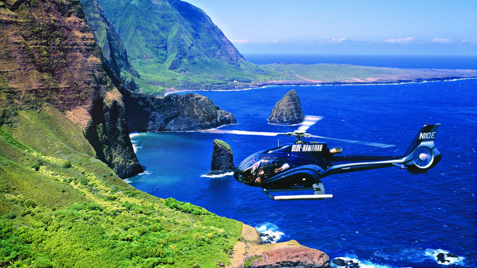 Helicopter flies over ocean and sweeping green cliffs