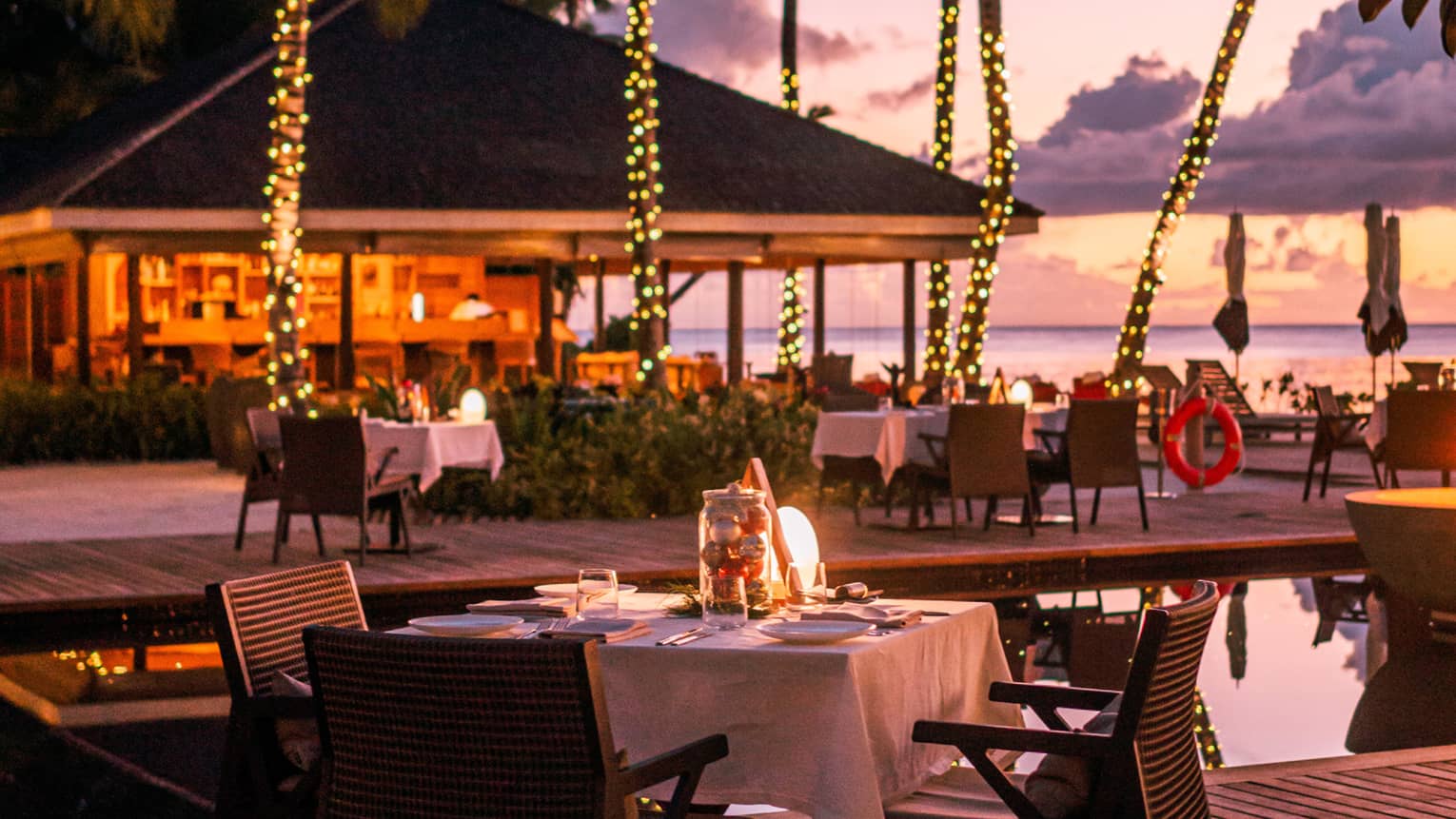 Claudine waterfront terrace with square dining tables amid palm trees wrapped in white lights at dusk