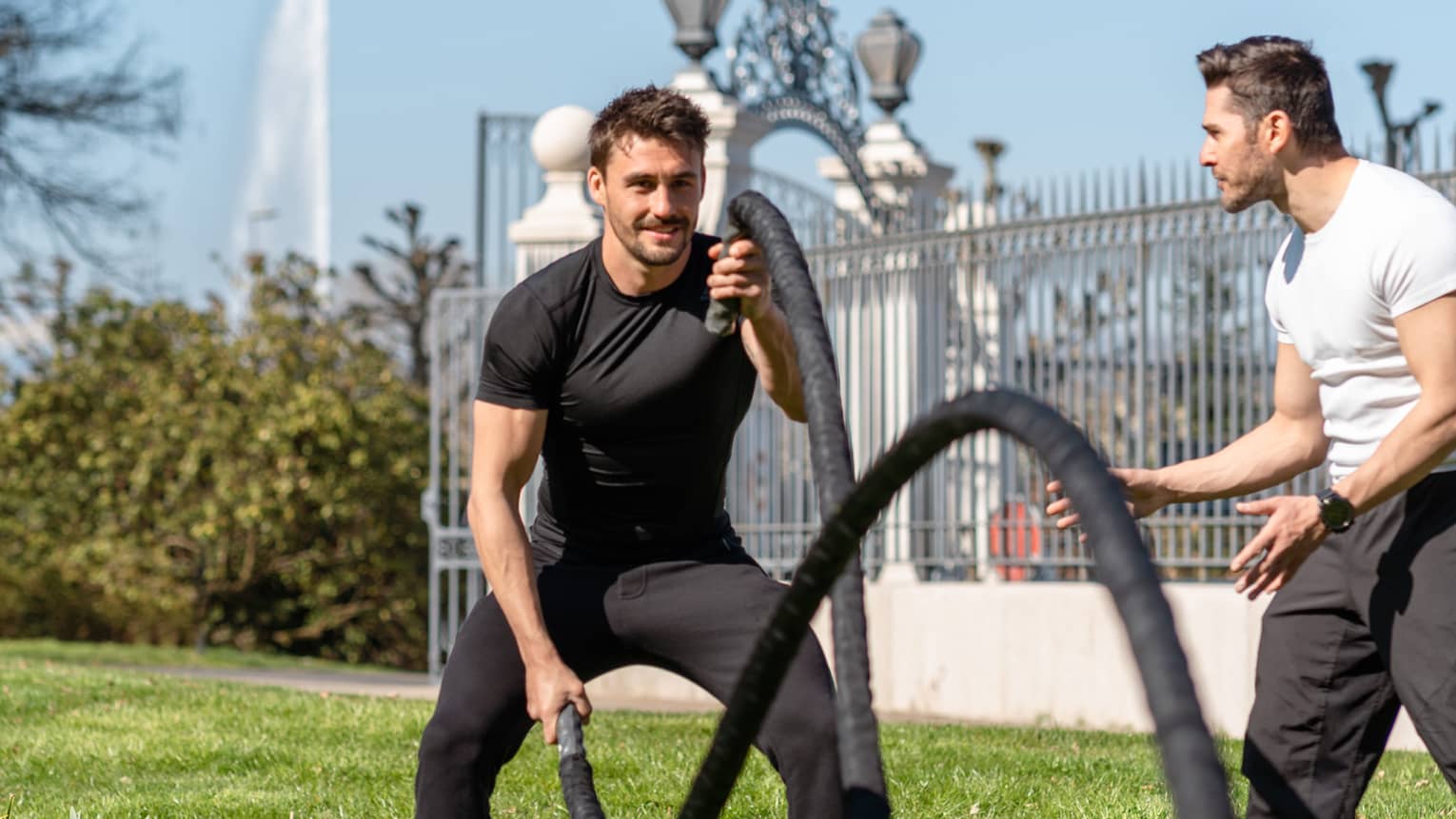 Man holds crossfit ropes as trainer watches on lawn in garden