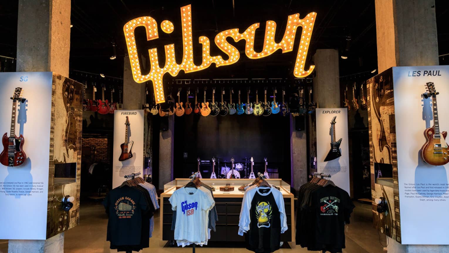 Interior of the Gibson Garage, featuring a large illuminated "Gibson" sign, displays of various guitar models, and racks of branded merchandise, with a stage in the background.