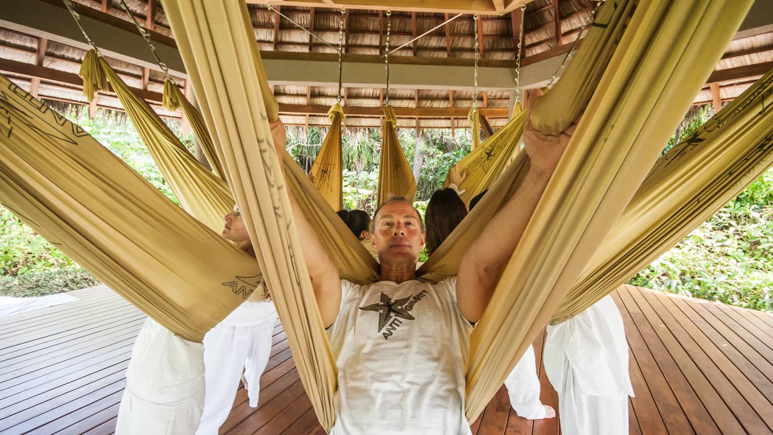 Group of people lie back into yellow slings attached to thatched roof during aerial yoga