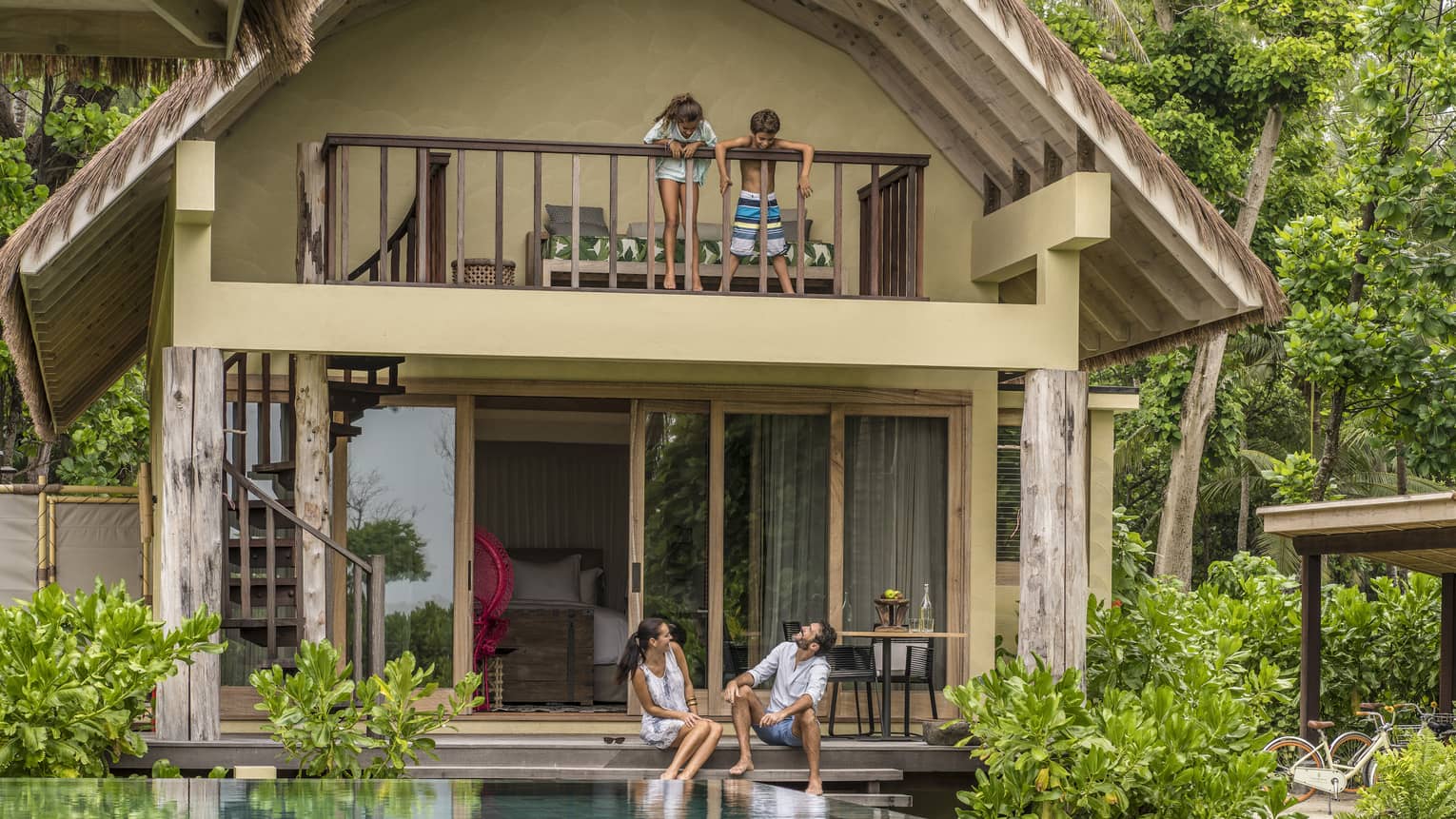 Couple with feet in private pool look up at two children standing on balcony of villa