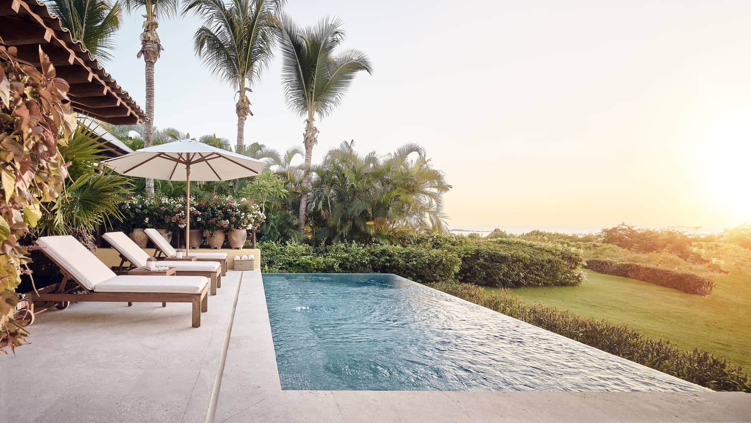 Three patio chairs, umbrella by long plunge pool overlooking lawn, sunset