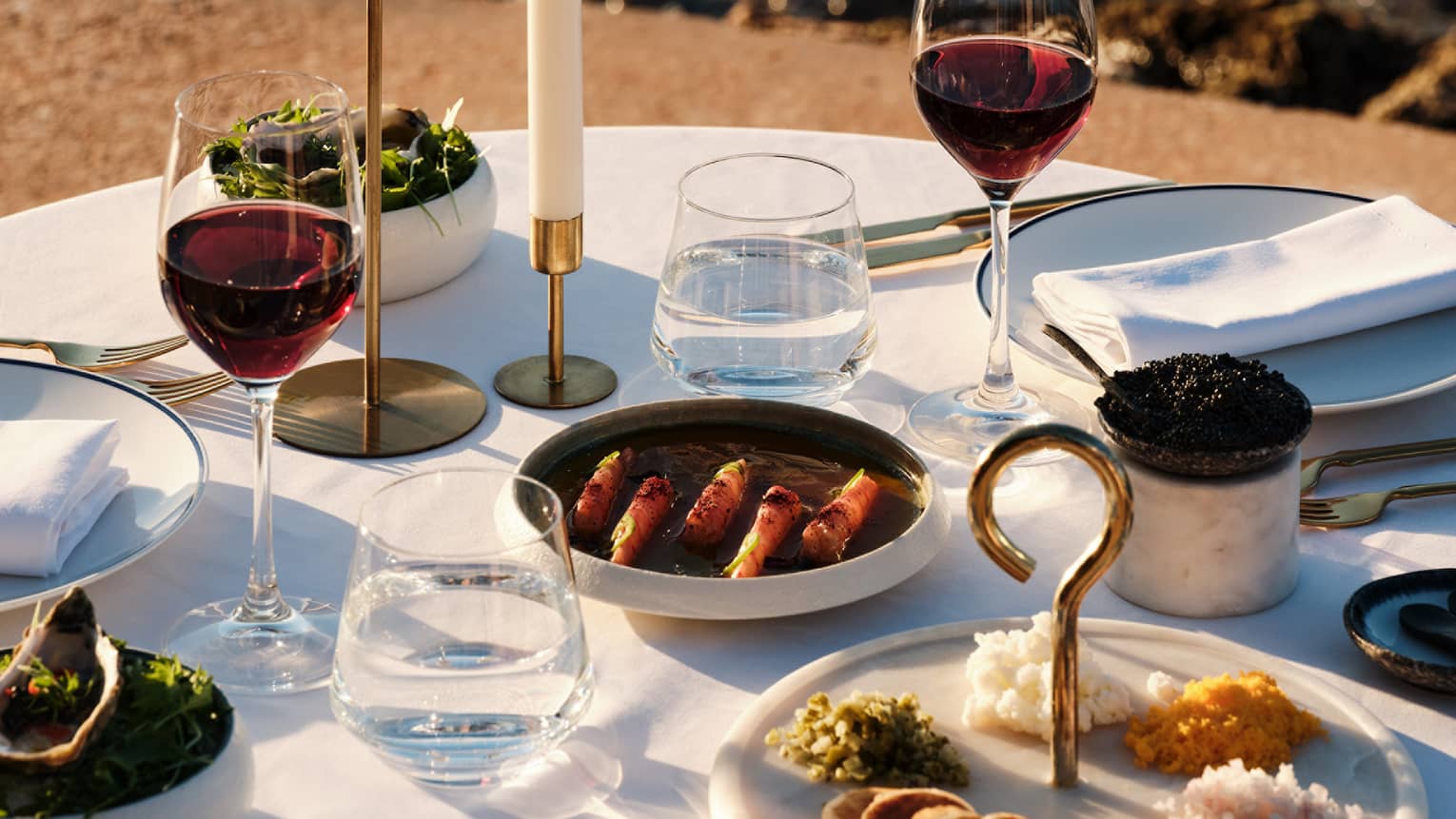 An elegant dining table in an outdoor setting. The table is set with candles, utensils, napkins, tumblers of water, wine glasses with wine, and dishes containing food items.