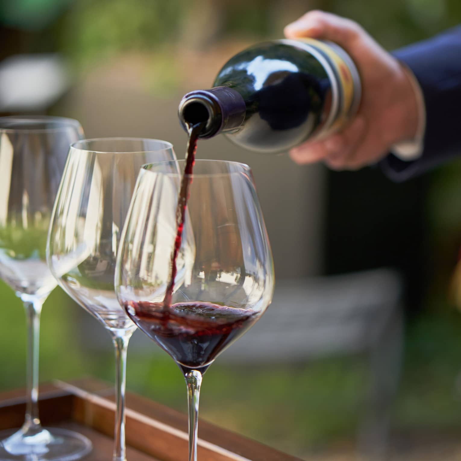 A close up of hands pouring a bottle of red wine into a clear wine glass that rests on a wooden tray, outdoors