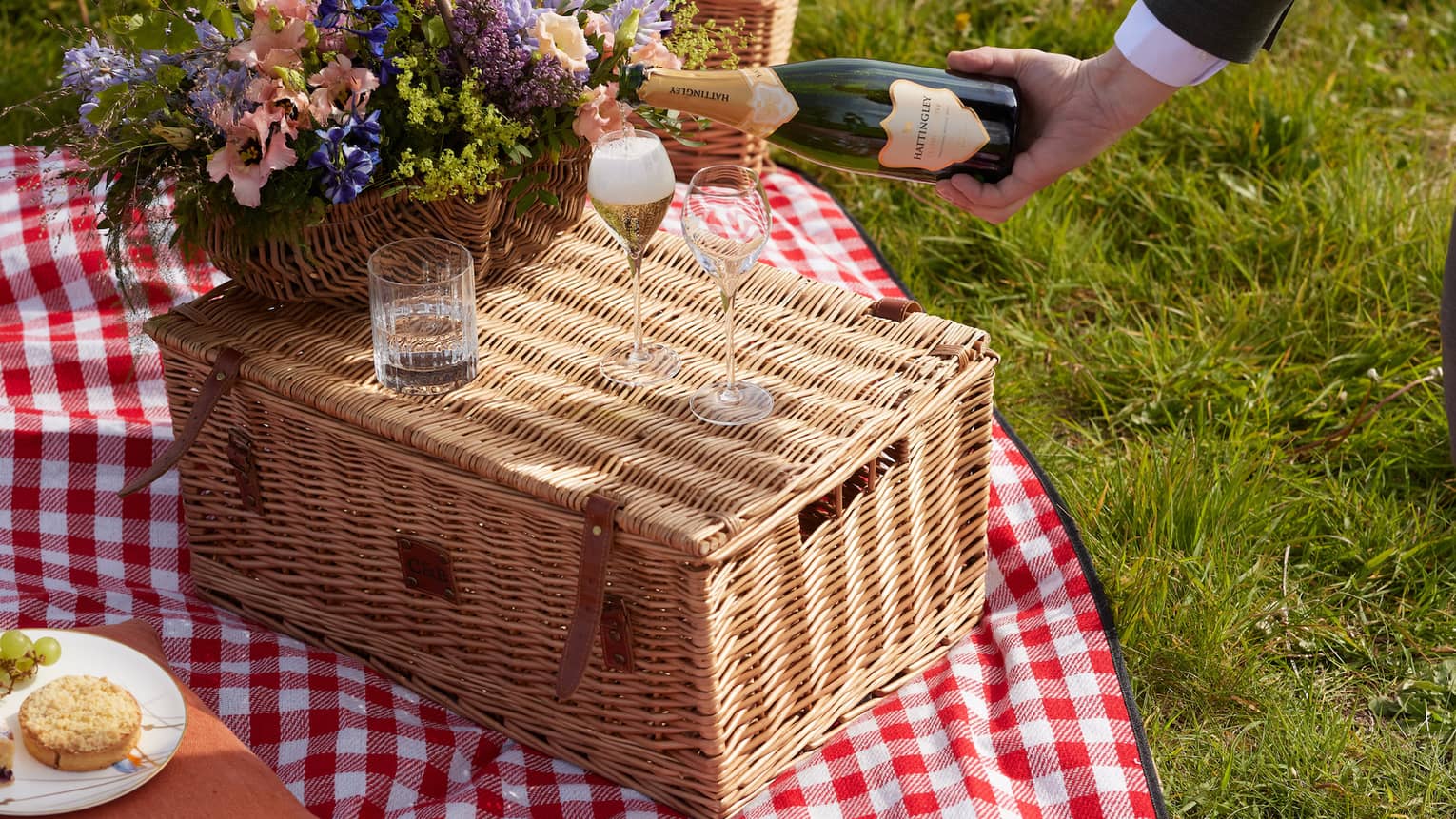 Attendant pours champagne into glass beside bouquet atop wicker picnic basket on a red-and-white gingham blanket