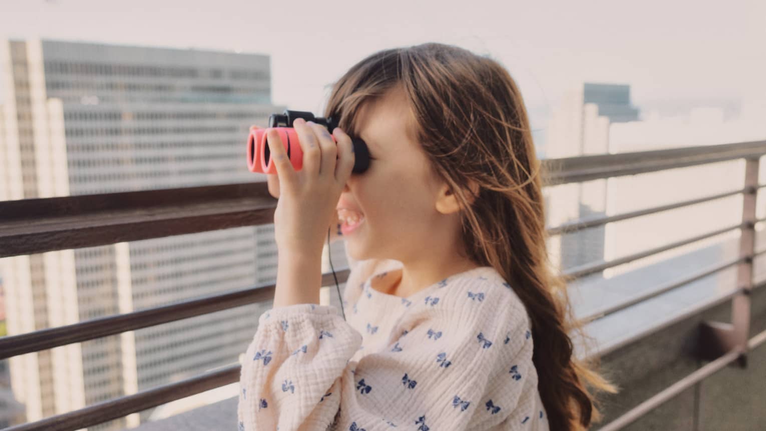 A little girl standing in front of a railing looks through pink binoculars with excitement.