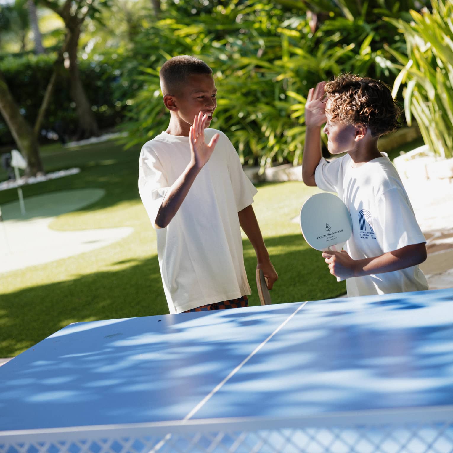 Two children high five each other while playing on an outdoor ping pong table