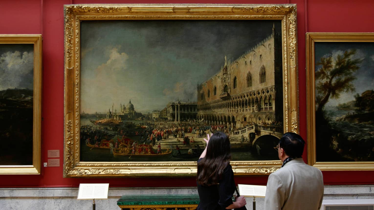 Back of two people looking up at large oil paintings with gold frames in Hermitage Museum