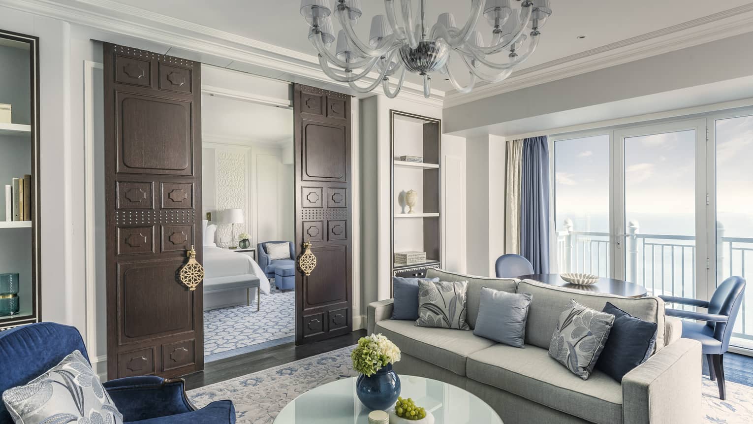 Sea-view living room, with grey sofa, blue pillows and accent chairs, chandelier, dark wooden doors open to bedroom