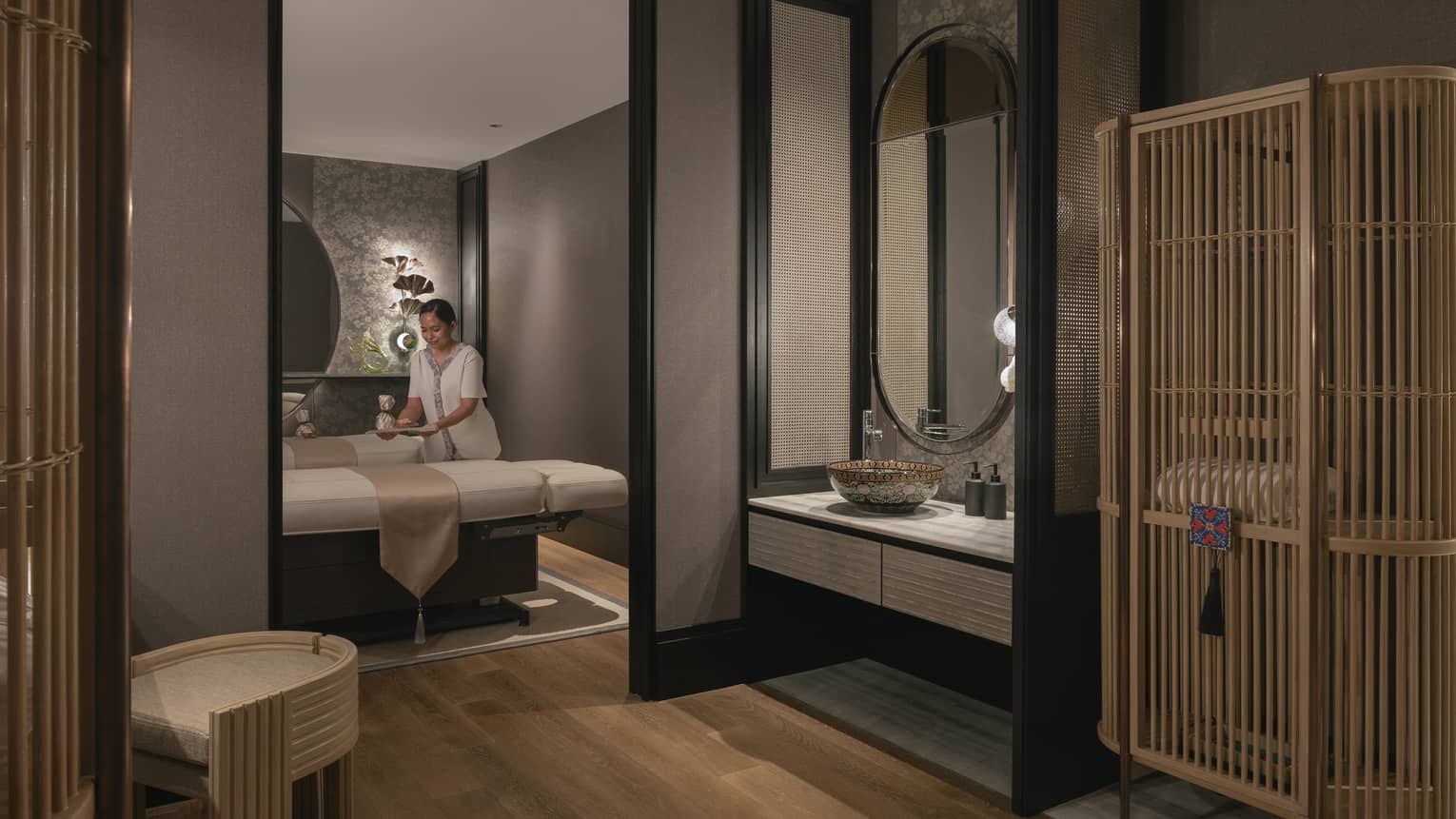 A tray is gently placed on one of two massage tables in a warmly lit spa room with soft grey walls and natural wood décor