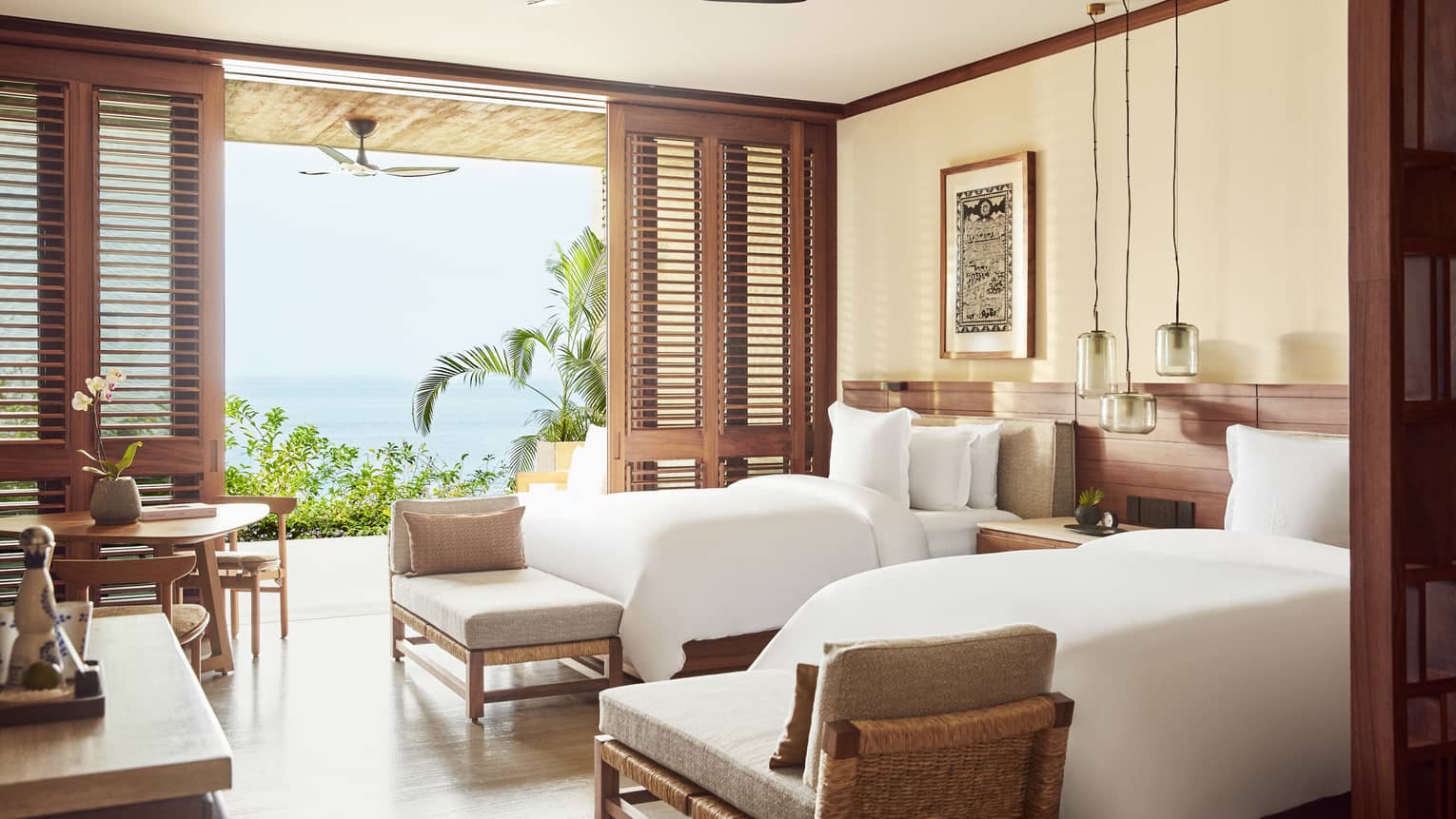 Bright bedroom with twin beds, white linens and wooden accents, opening to a terrace with ocean views. The room features a cozy seating area and hanging pendant lights.