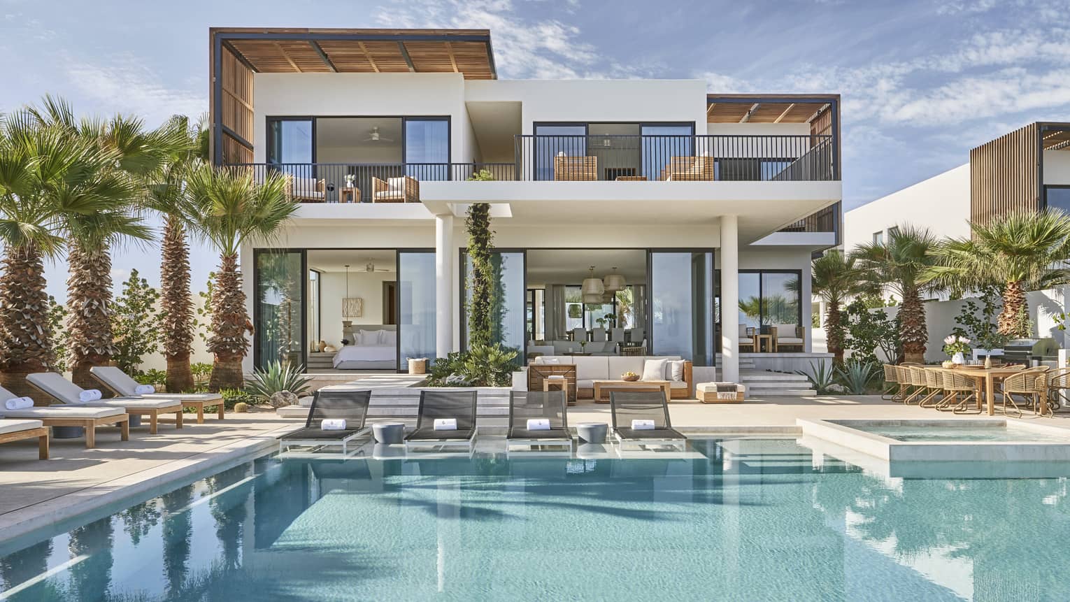 Modern luxury villa with a spacious pool area, surrounded by palm trees, featuring outdoor lounge chairs, a dining area, and floor-to-ceiling windows.
