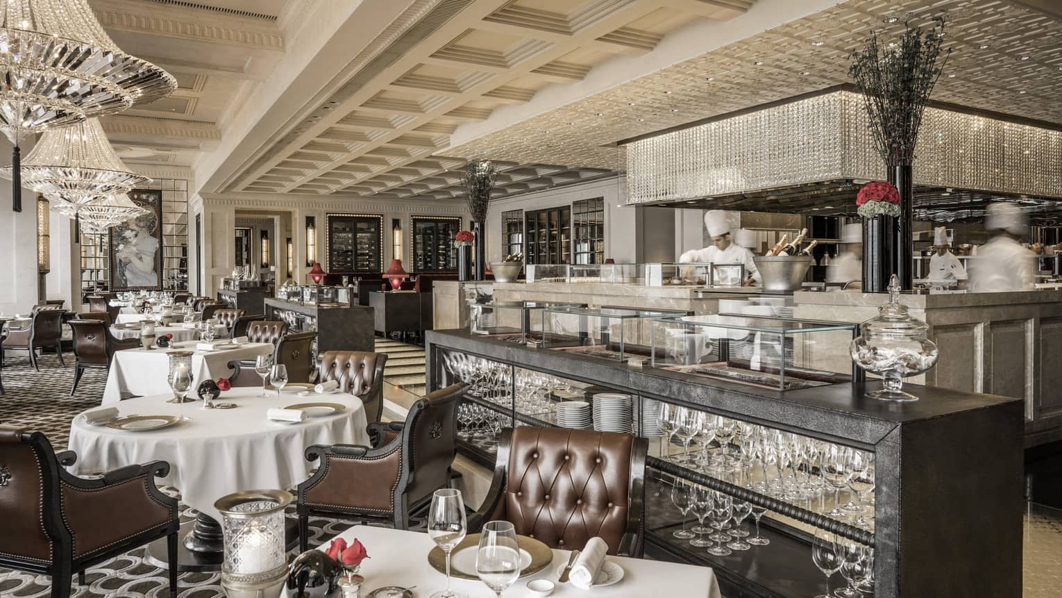 An elegant French restaurant interior with polished wooden floors, white tablecloth-covered tables, and plush leather chairs. The room features chandeliers. 