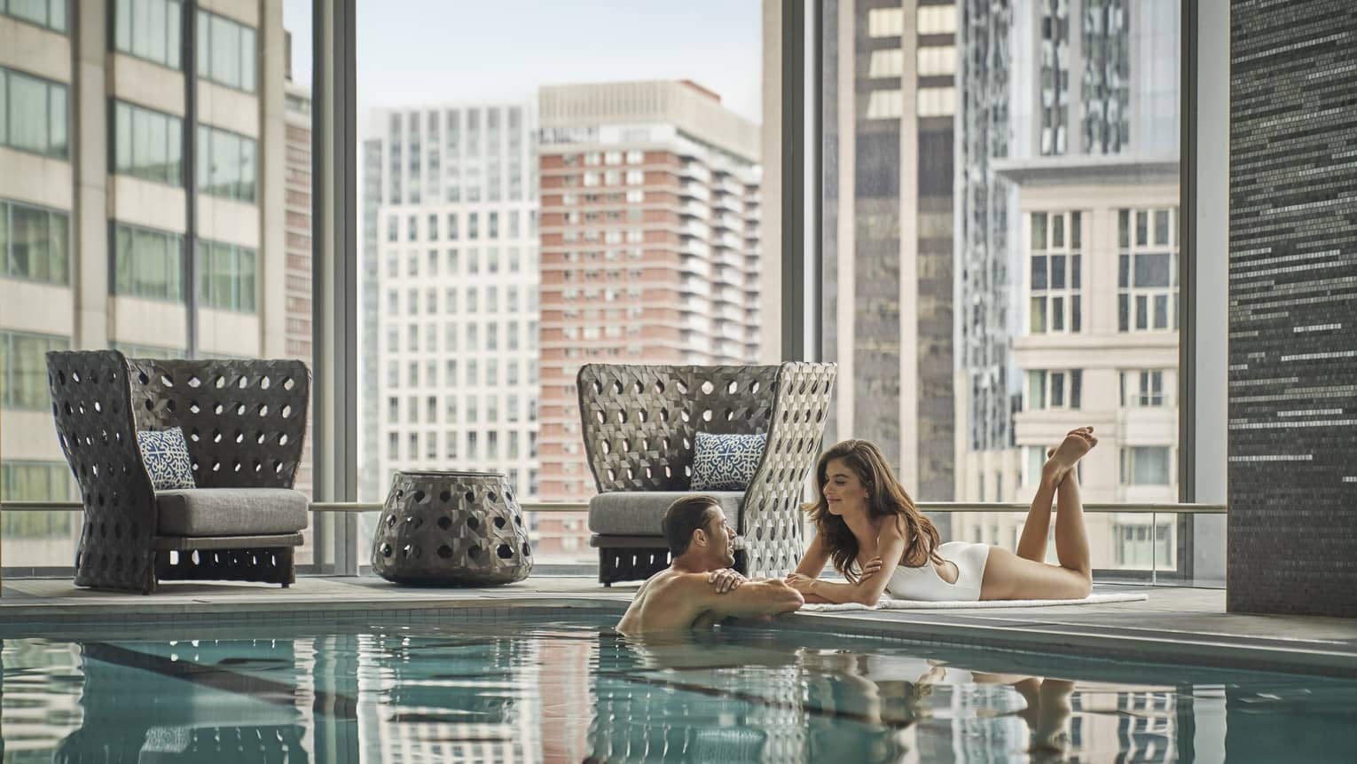 Man in indoor swimming pool looks up at woman in swimsuit lounging on deck