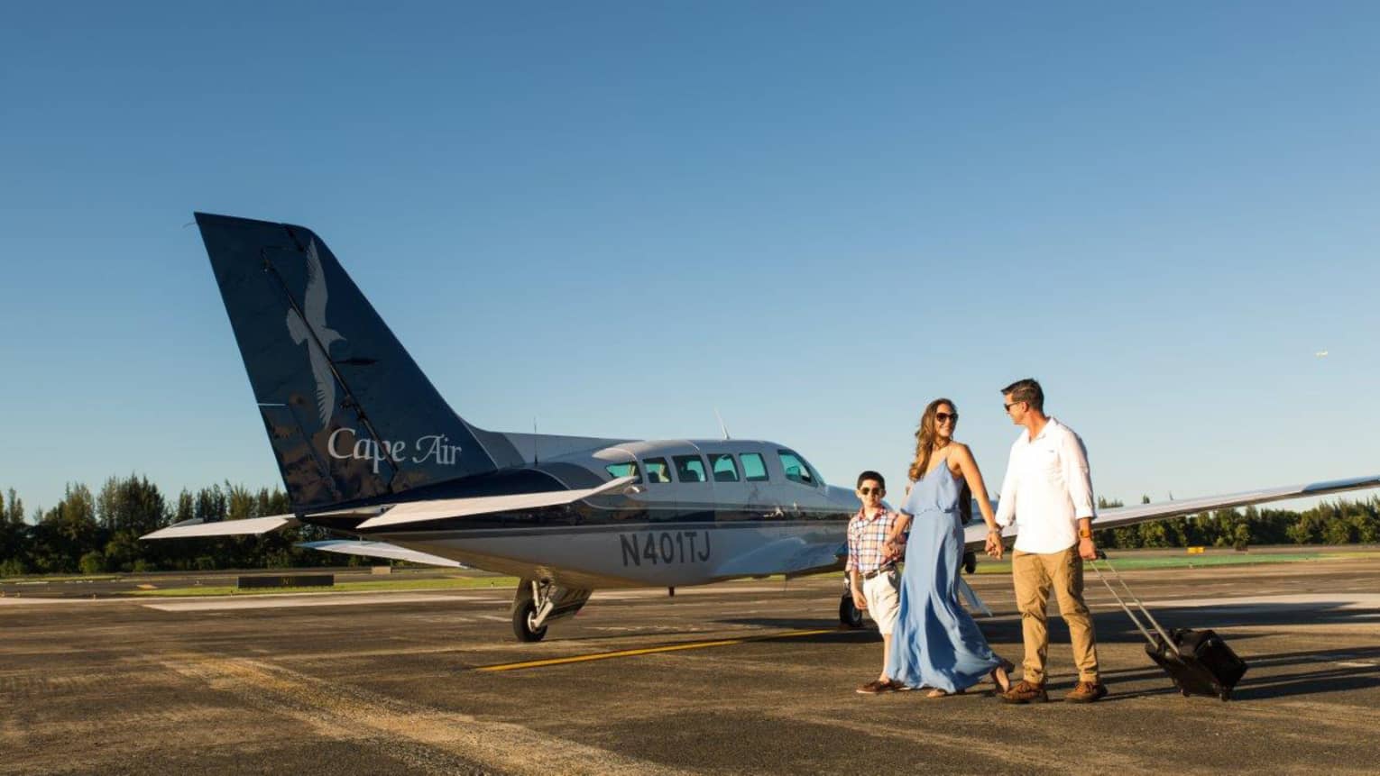 A mother, father and son walking away from a plane while the father pulls a suitcase.