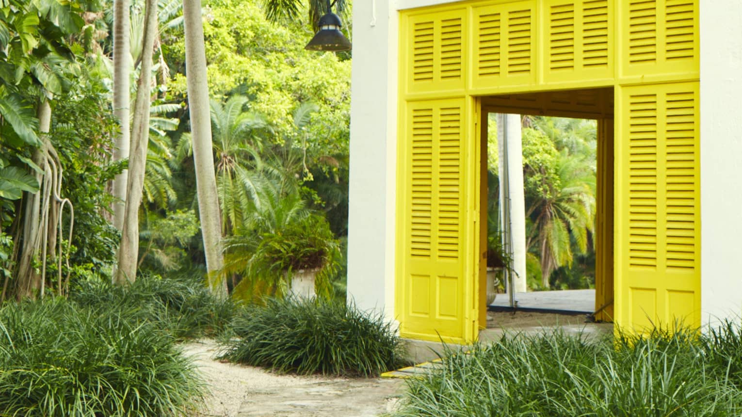 A cabana house with yellow shutter doors and a covered upper balcony with filigree railings is nestled amid lush foliage. 