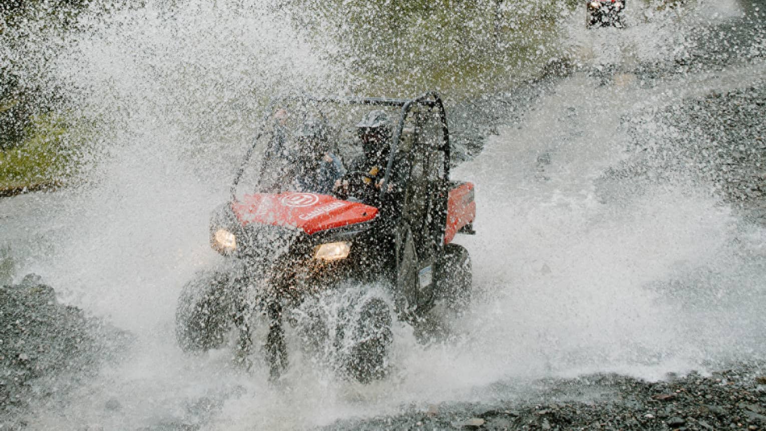 A red-and-black ATV driving through water, creating a thick veil of water spray against a background of pine forest.