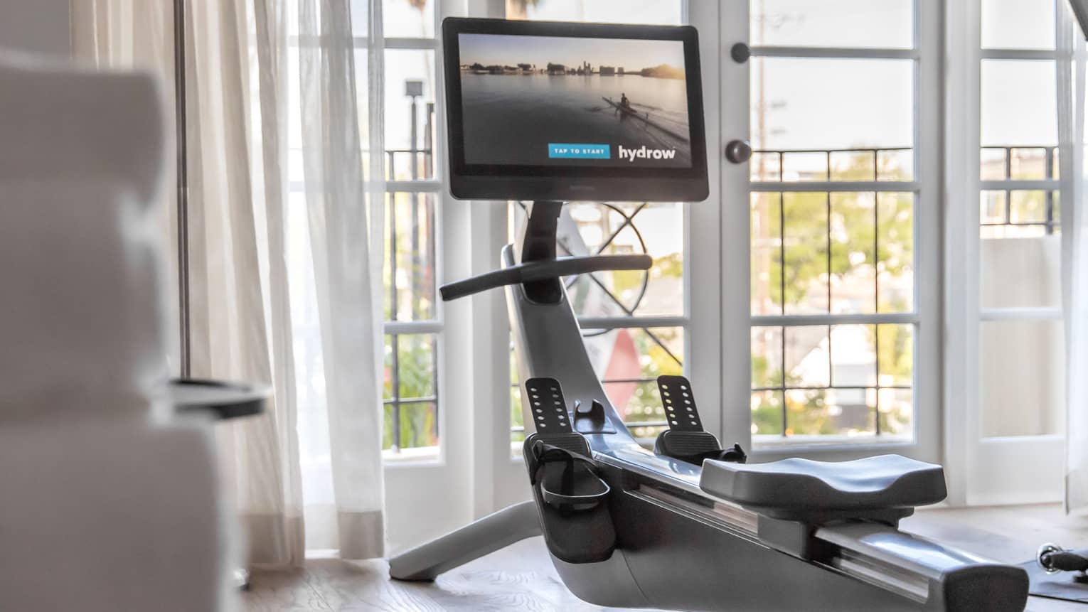 In a bright room, a rowing machine equipped with a screen faces two French doors, sheer white curtains pulled to the side.