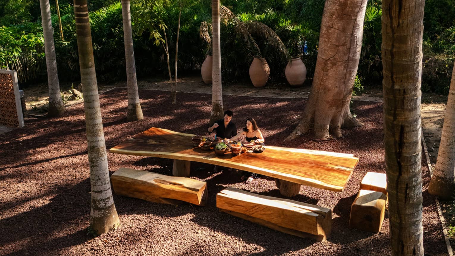Two people sit at a long wood outdoor table with wood beam bench seats, all surrounded by palm trees.