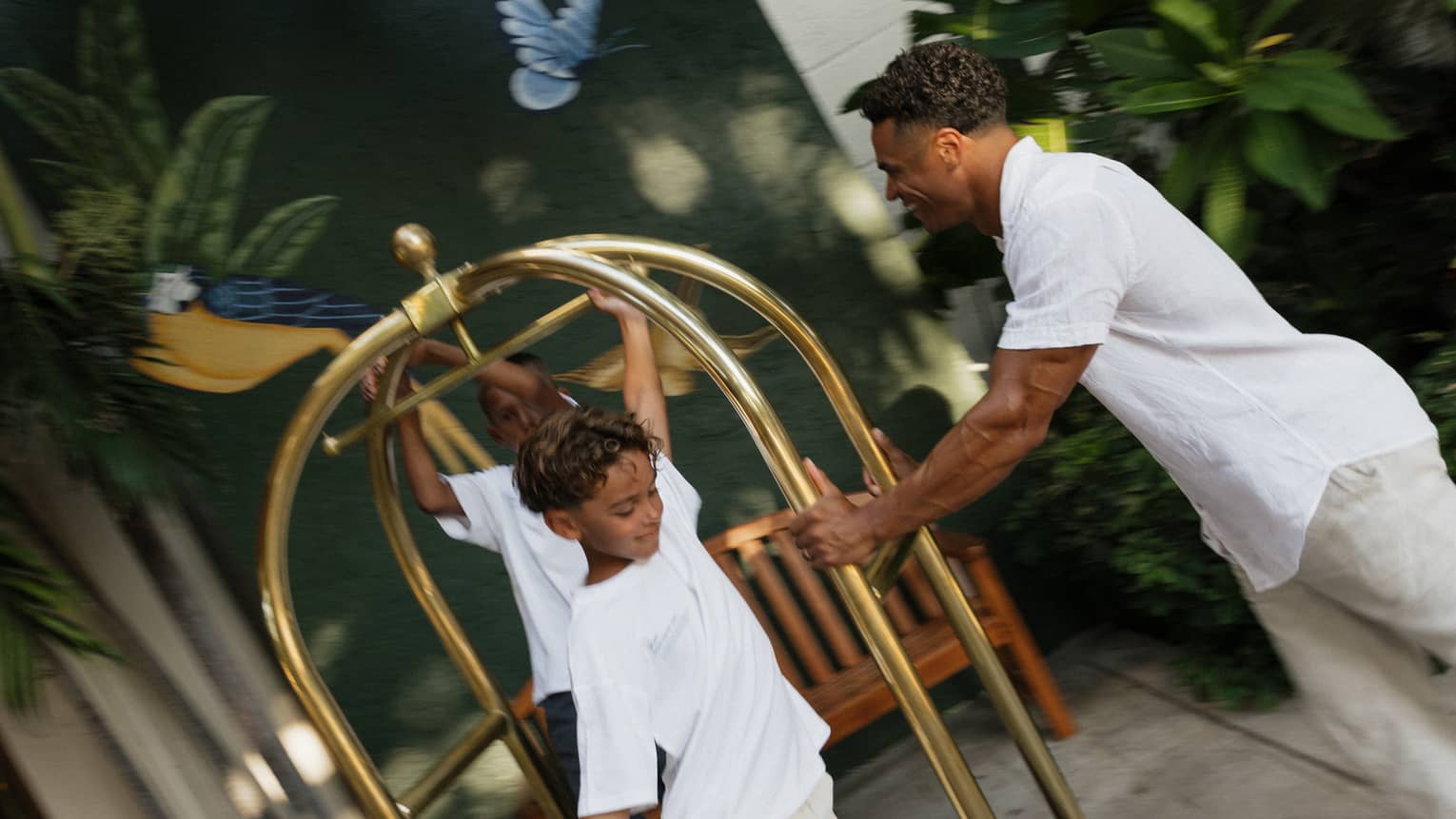 A man pushes two children on a gold luggage cart on a sidewalk
