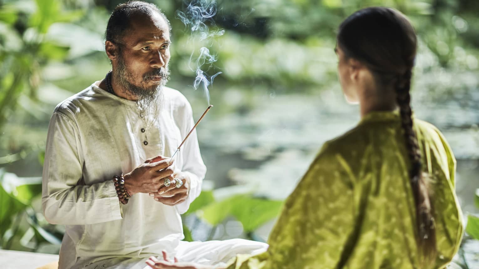 Set against a backdrop of greenery, Djik Dewa holds a stick of burning incense before a meditative cross-legged guest. 