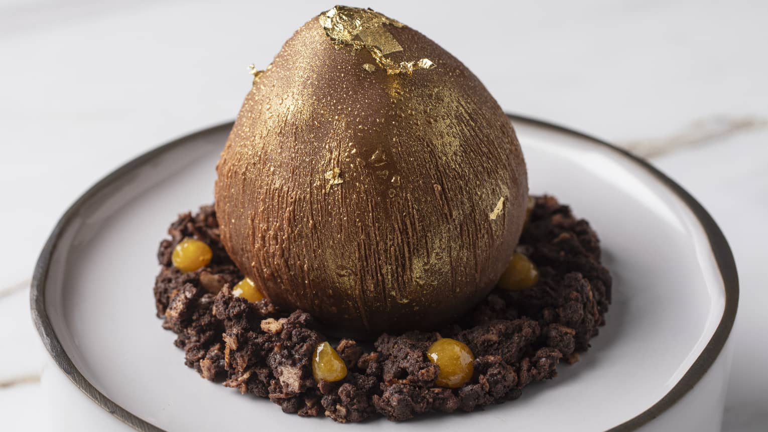 Dessert with caraibi mousse, caramel sauce, hazelnuts financier, hazelnuts crunchy, chocolate streusel and orange compote served on a white plate with a dark rim.