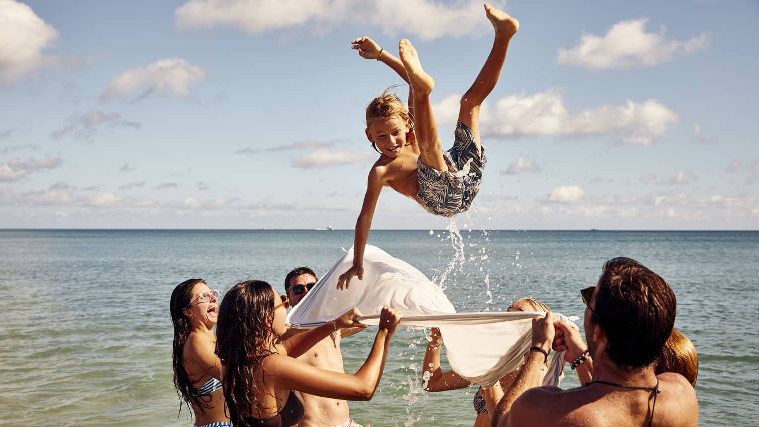 A group enjoys a sunny beach day, tossing a child into the air with a white sheet. The child smiles mid-air against clear skies.