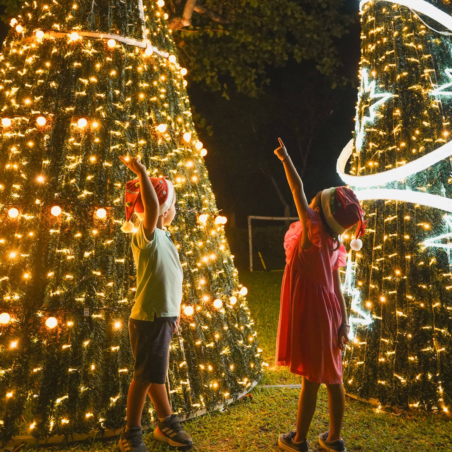 Two children wearing Santa hats, standing in front of two lit Christmas trees with one hand pointed upwards