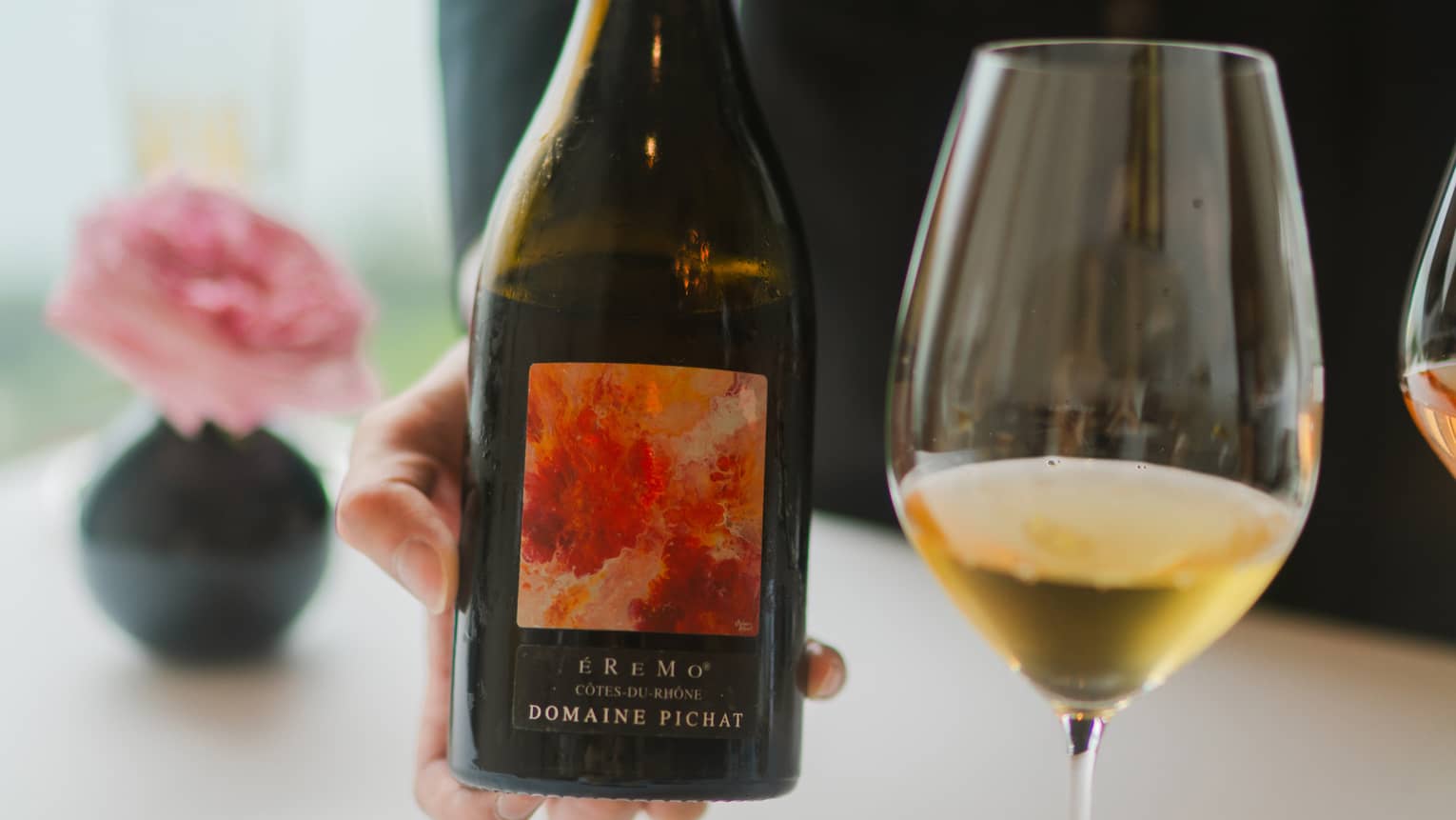 Person holding a wine bottle beside a glass of wine.