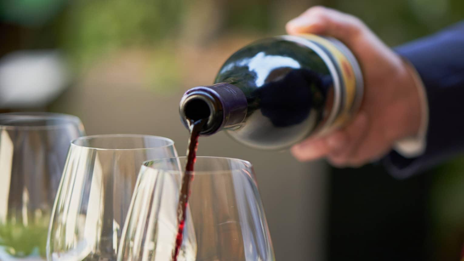 A close up of hands pouring a bottle of red wine into a clear wine glass that rests on a wooden tray, outdoors