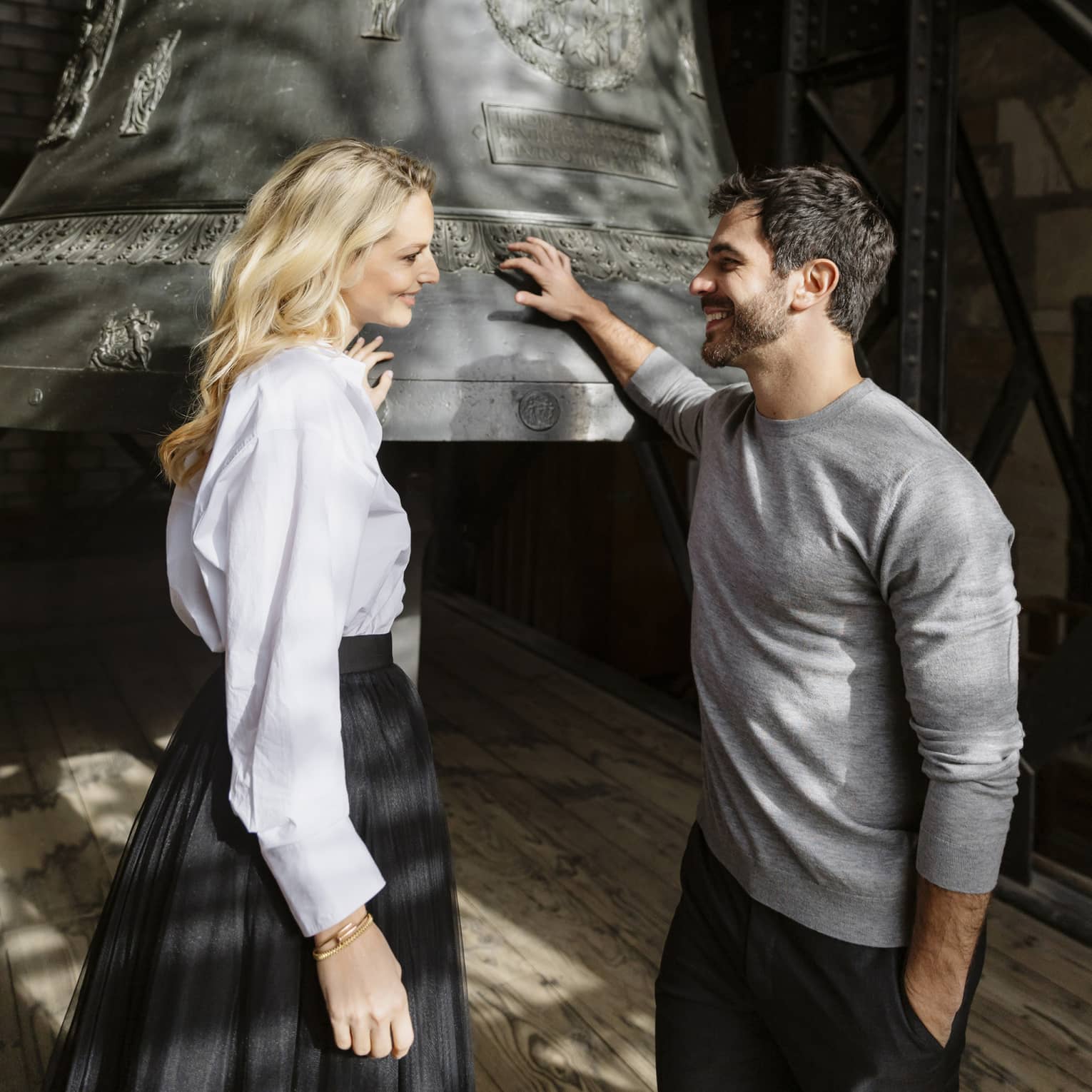 Two people stand next to a large metal bell in the bell tower of a cathedral.